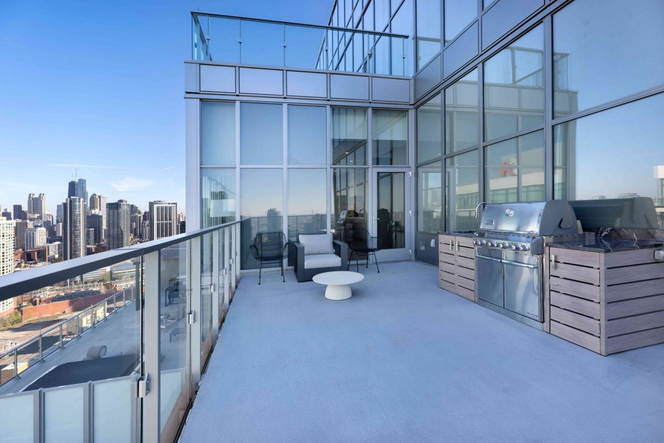 A modern rooftop terrace with glass railings, outdoor seating, a white round table, and a stainless steel grill, overlooking a city skyline under a clear blue sky.