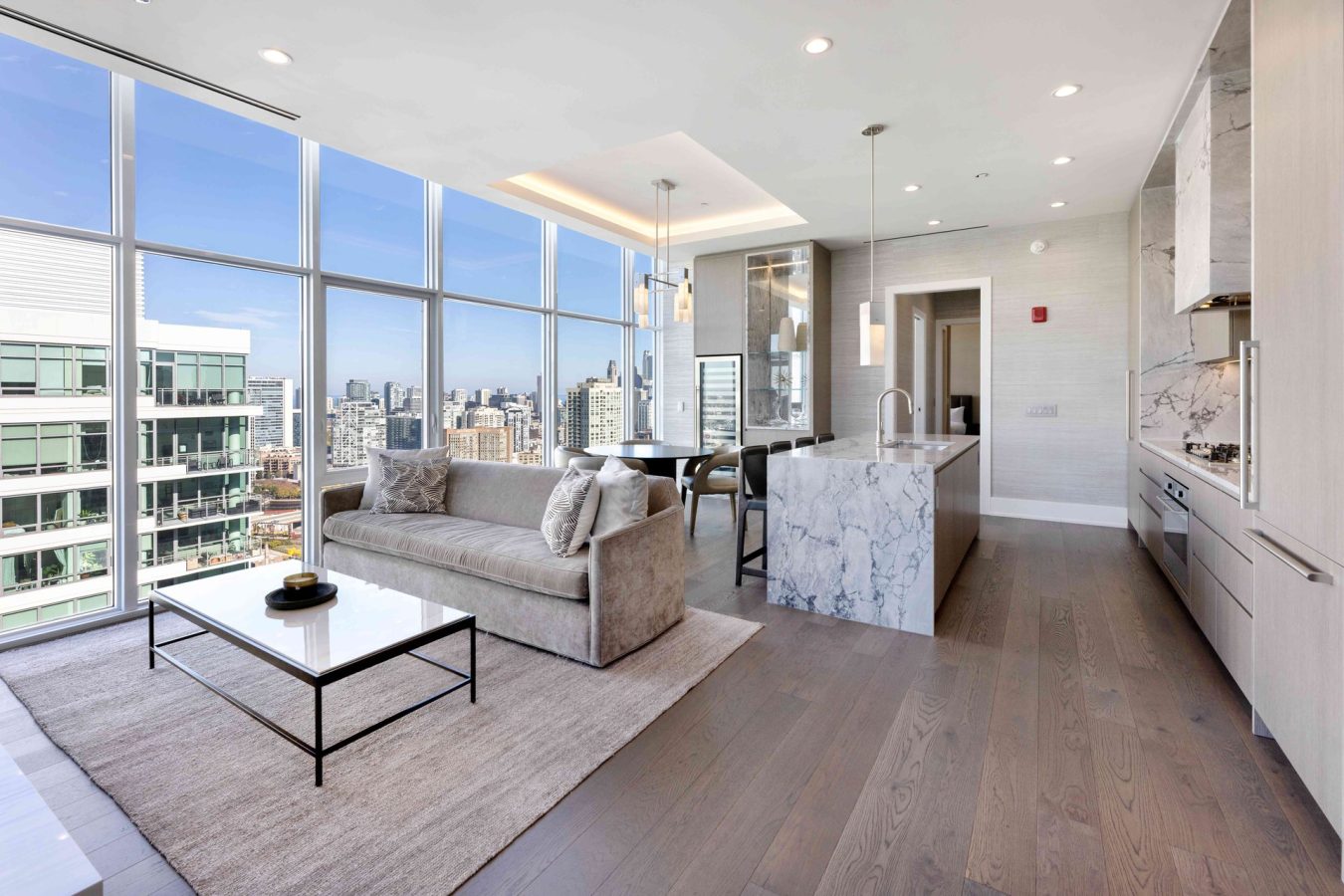 Modern high-rise apartment with floor-to-ceiling windows, city view, open-plan living area, beige sofa, marble kitchen island, built-in appliances, light wood flooring, and contemporary decor.