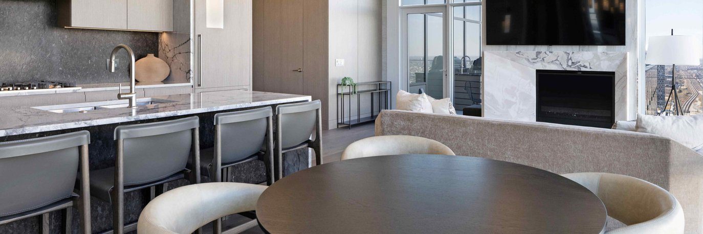 Modern, open-concept living space with a marble kitchen island and four barstools, round dining table with four chairs, light sofa, fireplace, large TV, and floor-to-ceiling windows overlooking a cityscape.