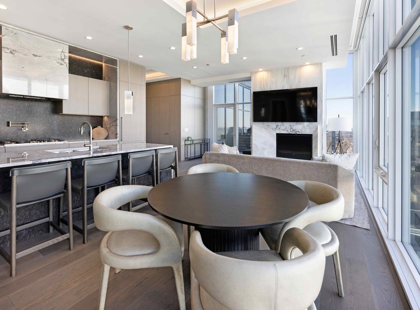 Modern open-concept kitchen and living room with floor-to-ceiling windows, round dining table with four chairs, marble countertops, barstools, sofa, fireplace, wall-mounted TV, and city skyline views.