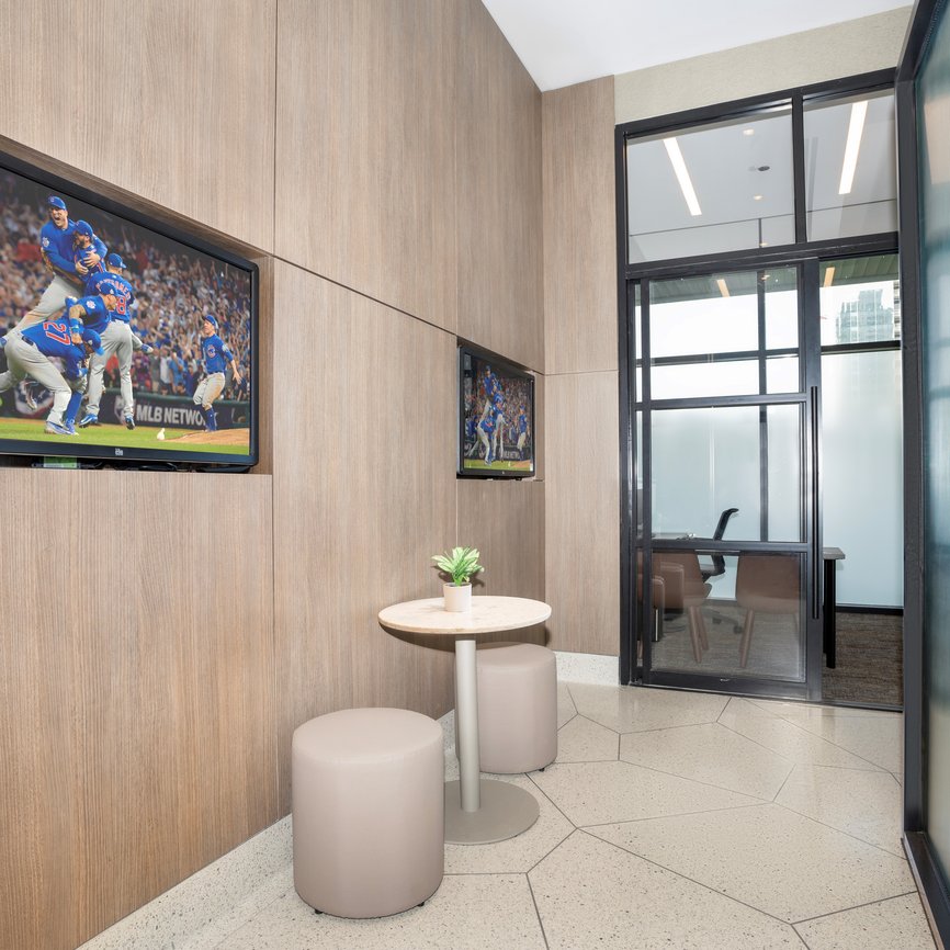 A modern lounge area with two round tables, beige stools, a small potted plant, and two wall-mounted TVs showing a baseball game. Glass doors lead to a bright office in the background.