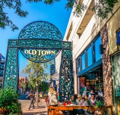 People sit at outdoor tables near a large decorative metal archway that reads Old Town on a sunny day, with shops and trees lining the street.