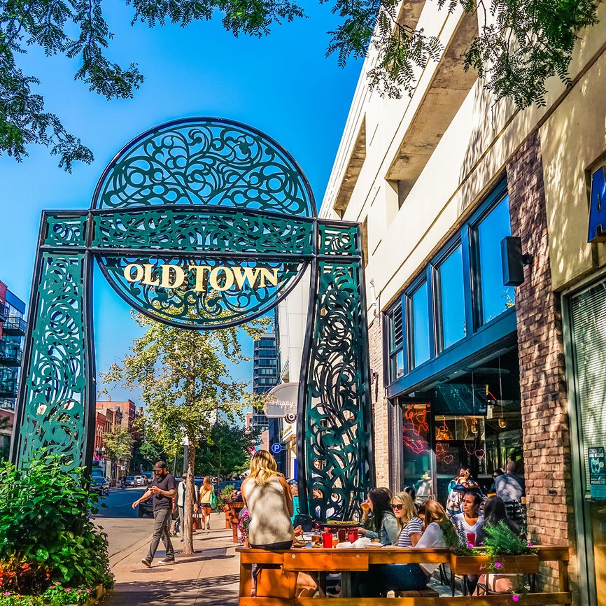 People sit at outdoor tables near a large decorative metal archway that reads Old Town on a sunny day, with shops and trees lining the street.