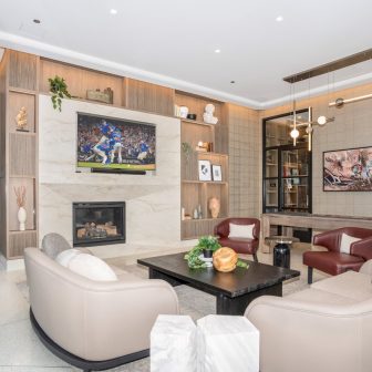 Modern living room with built-in shelves, a marble fireplace, a large TV showing a baseball game, tan and red chairs around a black coffee table, and decorative accents throughout the space.