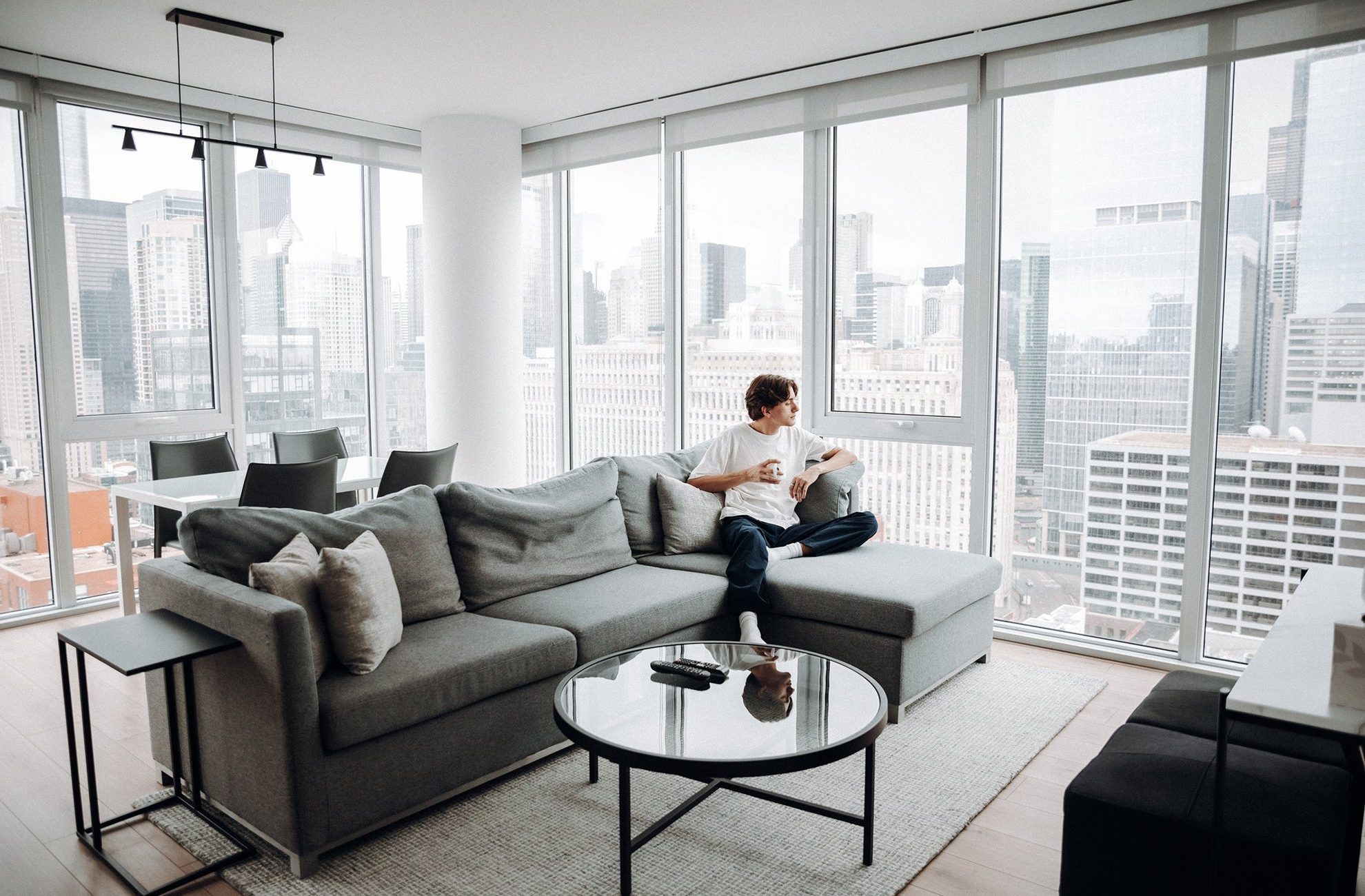 A person sits on a gray sectional sofa in a modern, bright apartment with floor-to-ceiling windows, holding a cup and looking out at the city skyline. A dining area is visible in the background.