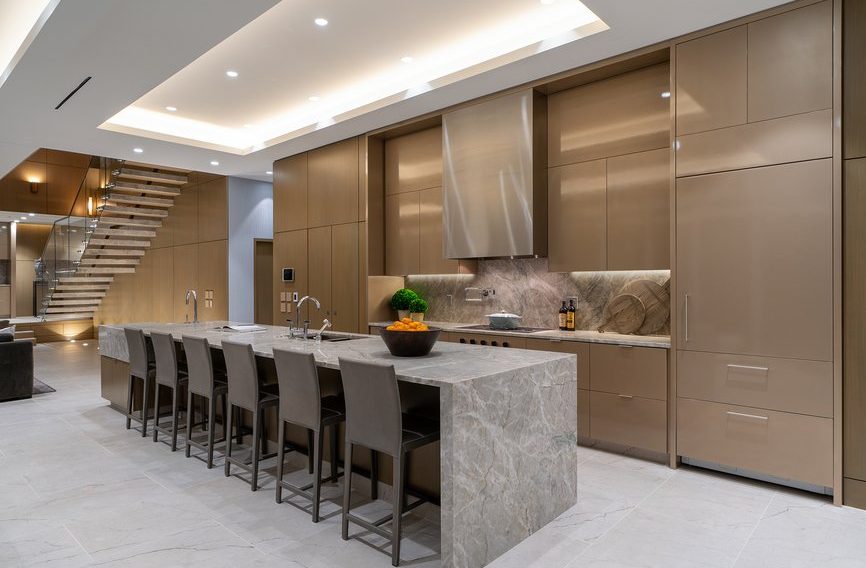 Modern kitchen with tan cabinetry, stainless steel appliances, a large gray marble island with six chairs, and a bowl of oranges on the countertop. Recessed lighting and an open staircase are visible in the background.