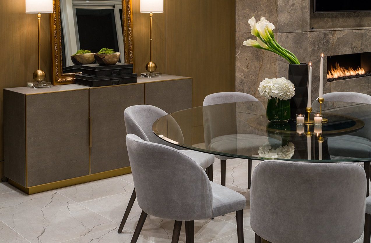 A modern dining room with a round glass table, gray velvet chairs, a sideboard with decor, a large mirror, lamps, flowers, and a marble fireplace. The setting is elegant and softly lit.
