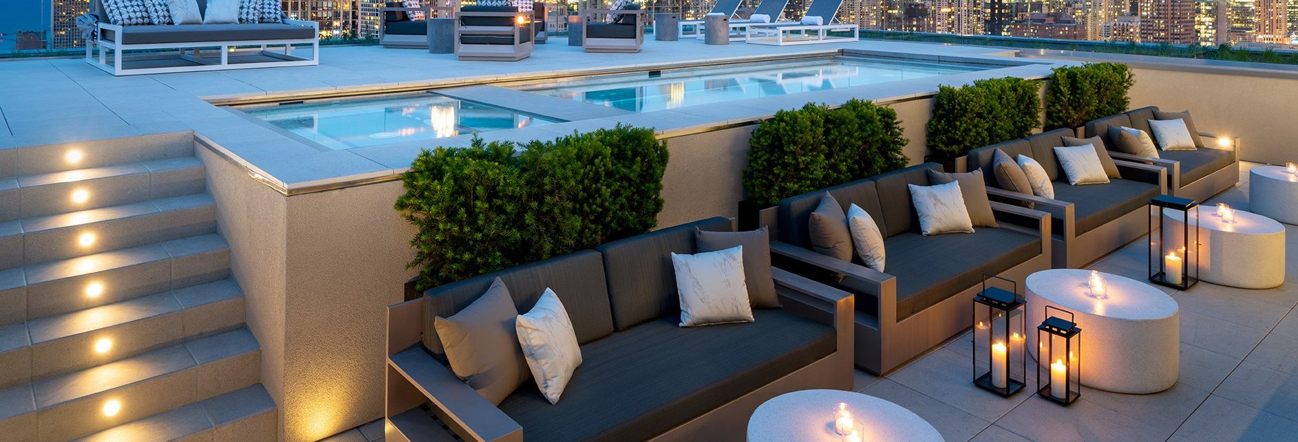 A modern rooftop patio at dusk features a lit swimming pool, lounge chairs, cushioned sofas with pillows, round tables with glowing candles, decorative lanterns, and city skyscrapers in the background.