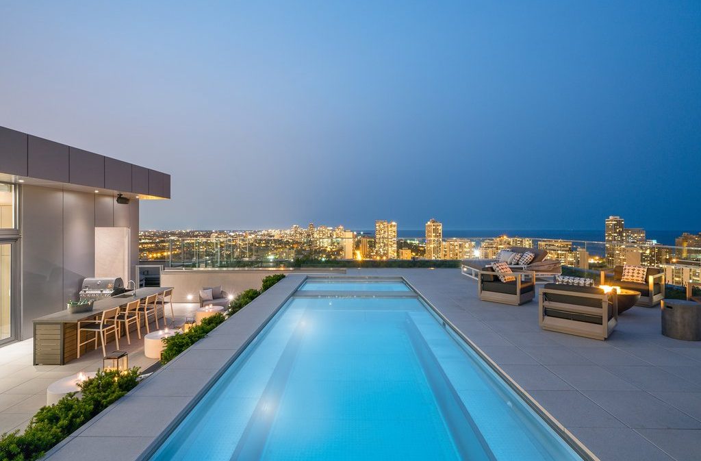 Rooftop patio at dusk with a lit swimming pool, outdoor kitchen, dining area, and lounge seating, overlooking a city skyline illuminated by building lights.