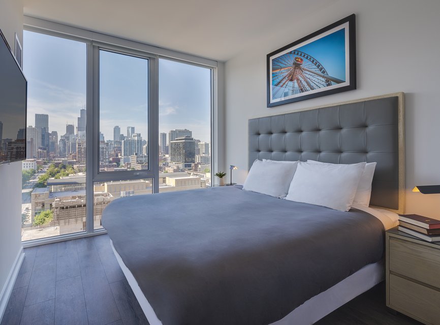 A modern bedroom with a large bed, gray headboard, and gray bedding, featuring floor-to-ceiling windows with a city skyline view. A framed ferris wheel photo hangs above the bed.