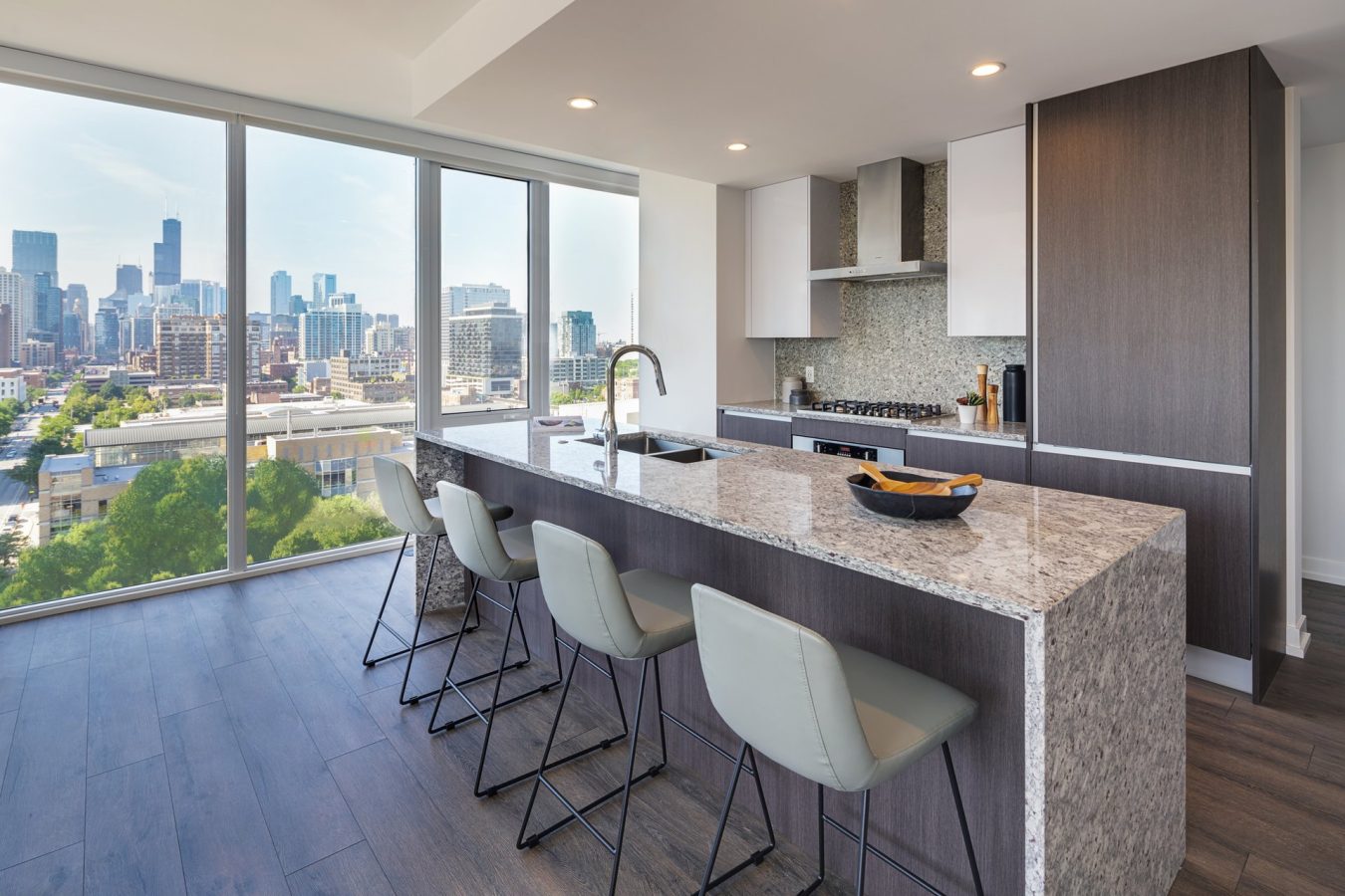 Modern kitchen with a large marble island, four barstools, and floor-to-ceiling windows offering a city skyline view. The kitchen features sleek cabinetry, built-in appliances, and wood flooring.