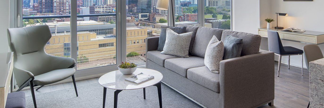 A modern living room with a gray sofa, armchair, coffee table, and floor-to-ceiling windows overlooking a cityscape. A desk with a chair and lamp is set up in the corner near the window.