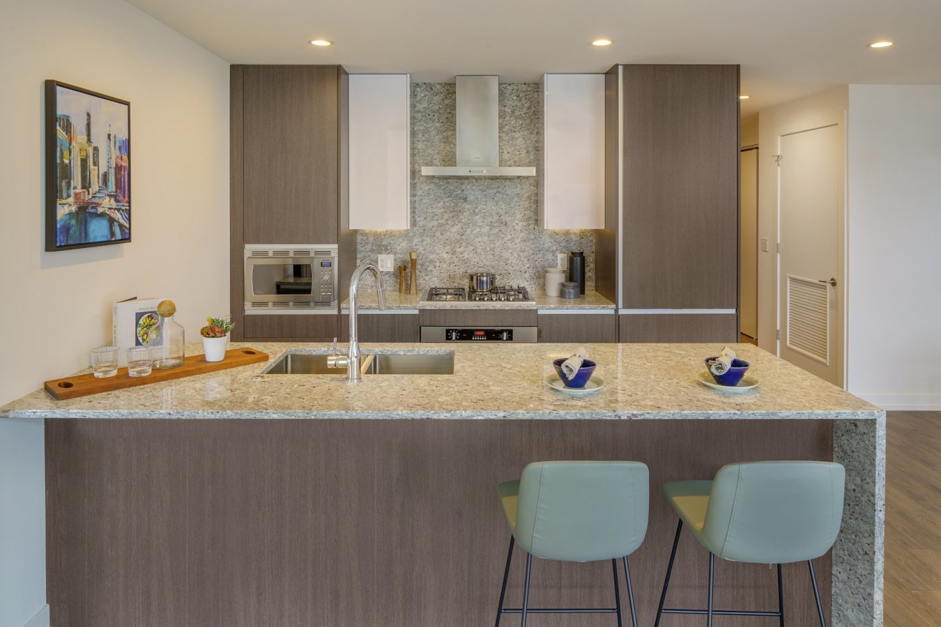 Modern kitchen with a marble island, two green bar stools, stainless steel appliances, wood cabinets, a built-in oven, and a small painting on the wall. The countertop has bowls, glasses, and a potted plant.