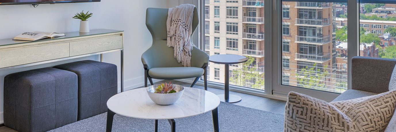 Modern living room with a gray chair draped with a blanket, round white coffee table with a plant, side table, two ottomans under a console, and large windows overlooking city buildings.