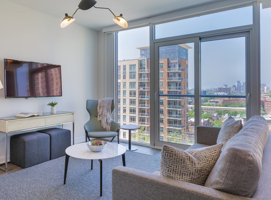 Modern living room with large windows, a gray sofa, patterned pillow, armchair with a throw, wall-mounted TV, and a white coffee table. City buildings and skyline visible outside. Bright, airy, and tidy space.