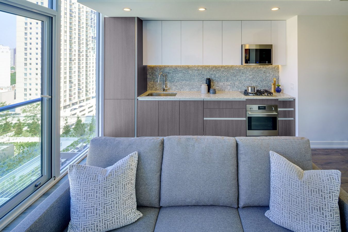 Modern apartment kitchen with light wood cabinets, a marble backsplash, stainless steel appliances, and a gray sofa with patterned pillows in the foreground. Large window shows city buildings and green park outside.