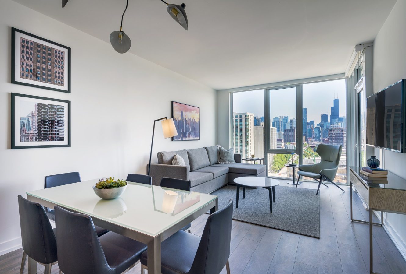 Modern apartment living room with a sofa, coffee table, dining table with chairs, large windows offering a city skyline view, wall-mounted TV, and artwork on white walls. Natural light fills the space.