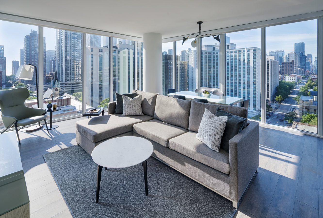 Modern living room with a large gray sectional sofa, round white coffee table, and floor-to-ceiling windows offering a city skyline view. Dining table and contemporary decor are visible, with sunlight streaming into the space.