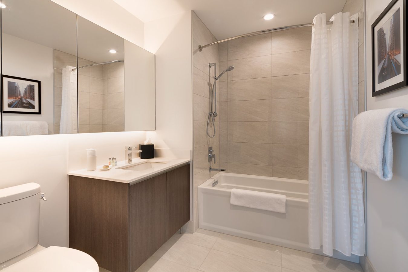 Modern bathroom with a large mirror, wooden vanity, toilet, bathtub with shower, tiled walls, white shower curtain, and a towel hanging on the wall. Decor includes framed art and toiletries on the counter.