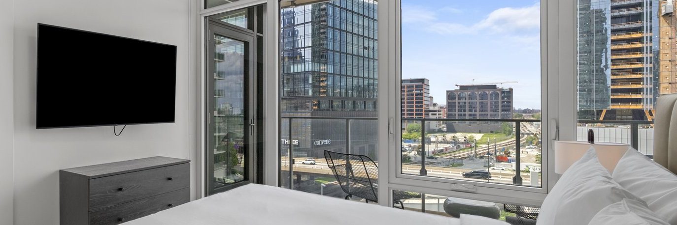 Modern bedroom with large windows showing a cityscape with glass buildings and construction. A bed with white linens and pillows is in the foreground, and a flat-screen TV is mounted on the wall next to a dresser.