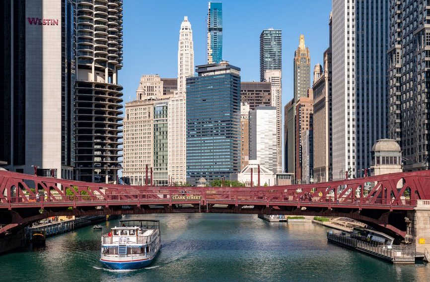A boat travels under a red bridge on a river lined with tall modern and historic skyscrapers in downtown Chicago on a clear, sunny day.