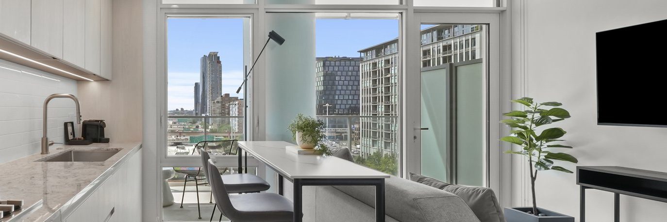 Modern apartment interior with a kitchen on the left, a dining table and chairs in the center, a sofa, potted plants, and large windows revealing a cityscape with tall buildings outside.