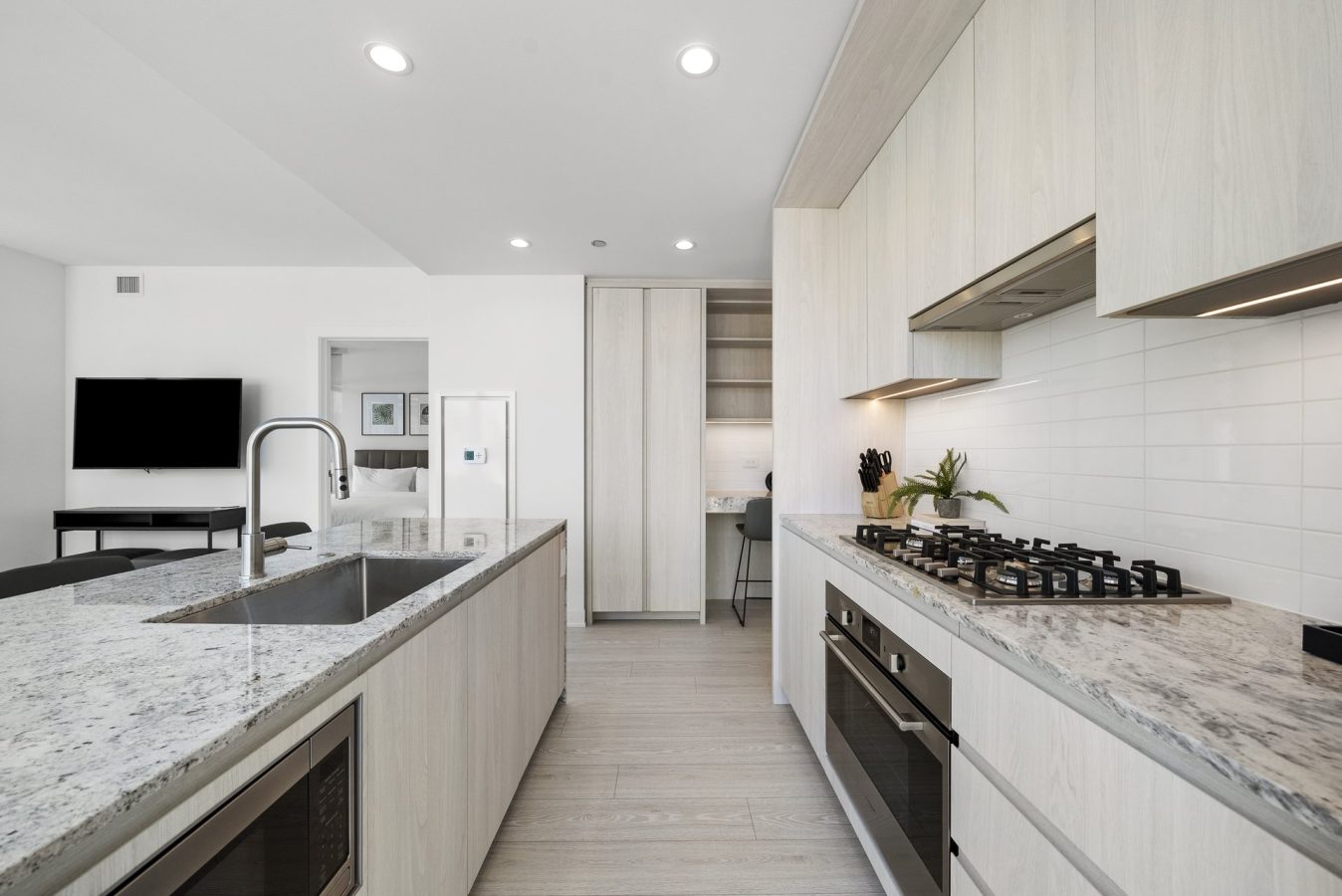 Modern kitchen with light wood cabinets, marble countertops, built-in oven, gas stovetop, stainless steel sink, and an island. In the background, a living area with a TV and a small workspace are visible.