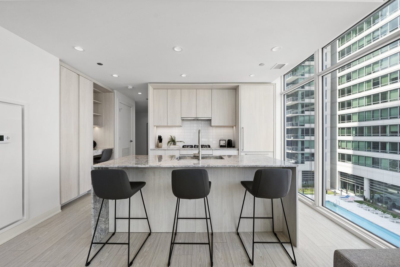 Modern kitchen with light wood cabinets, a marble island with three black barstools, and large floor-to-ceiling windows overlooking tall buildings and a pool outside.