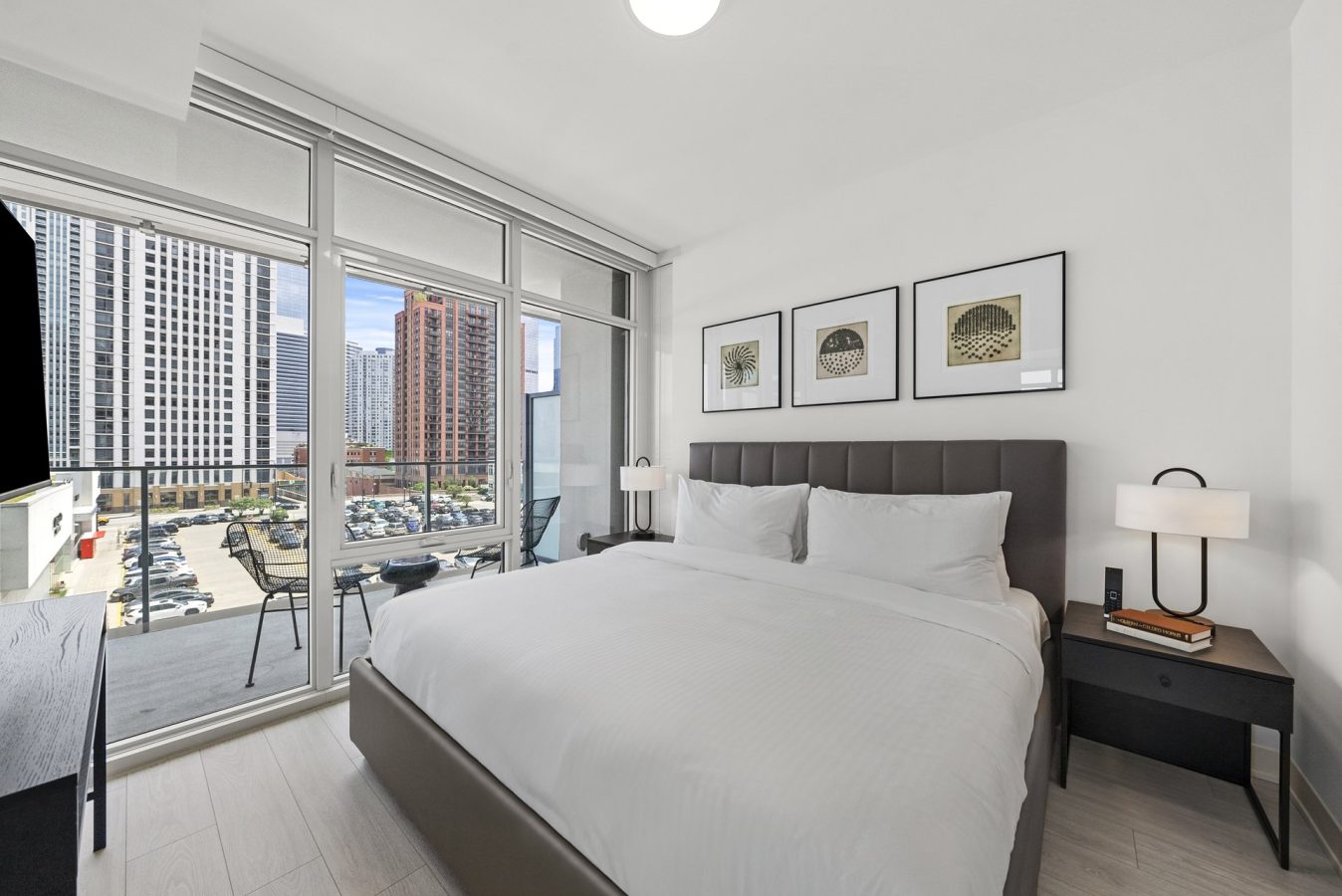 Modern bedroom with large bed, nightstands, and lamps; three framed artworks above the bed; floor-to-ceiling windows open to a balcony with chairs, overlooking tall city buildings and a parking lot.