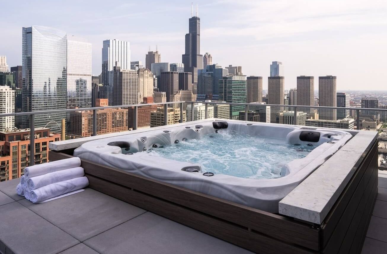 A rooftop hot tub with bubbling water and three stacked towels overlooks a city skyline with tall modern buildings under a clear sky.