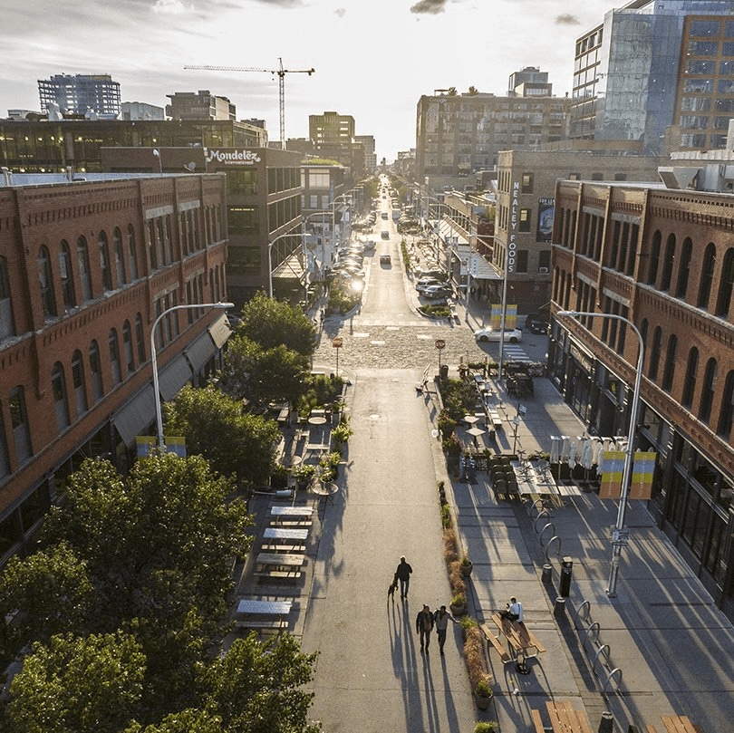 Aerial view of a city street lined with brick buildings, outdoor seating, and trees; a few people walk down the street, and sunlight casts long shadows across the scene.