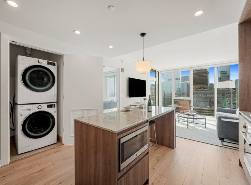 Modern apartment kitchen with a marble island, built-in microwave, and in-unit washer and dryer. The space opens to a living area with large windows showcasing a city skyline view.