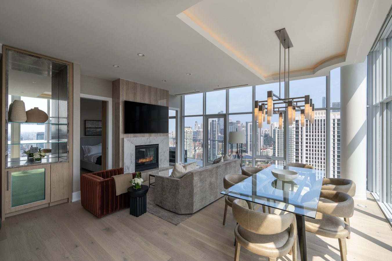 A modern high-rise apartment living and dining area with large windows offering a city view, neutral-toned furniture, a glass dining table, and a fireplace beneath a wall-mounted TV.