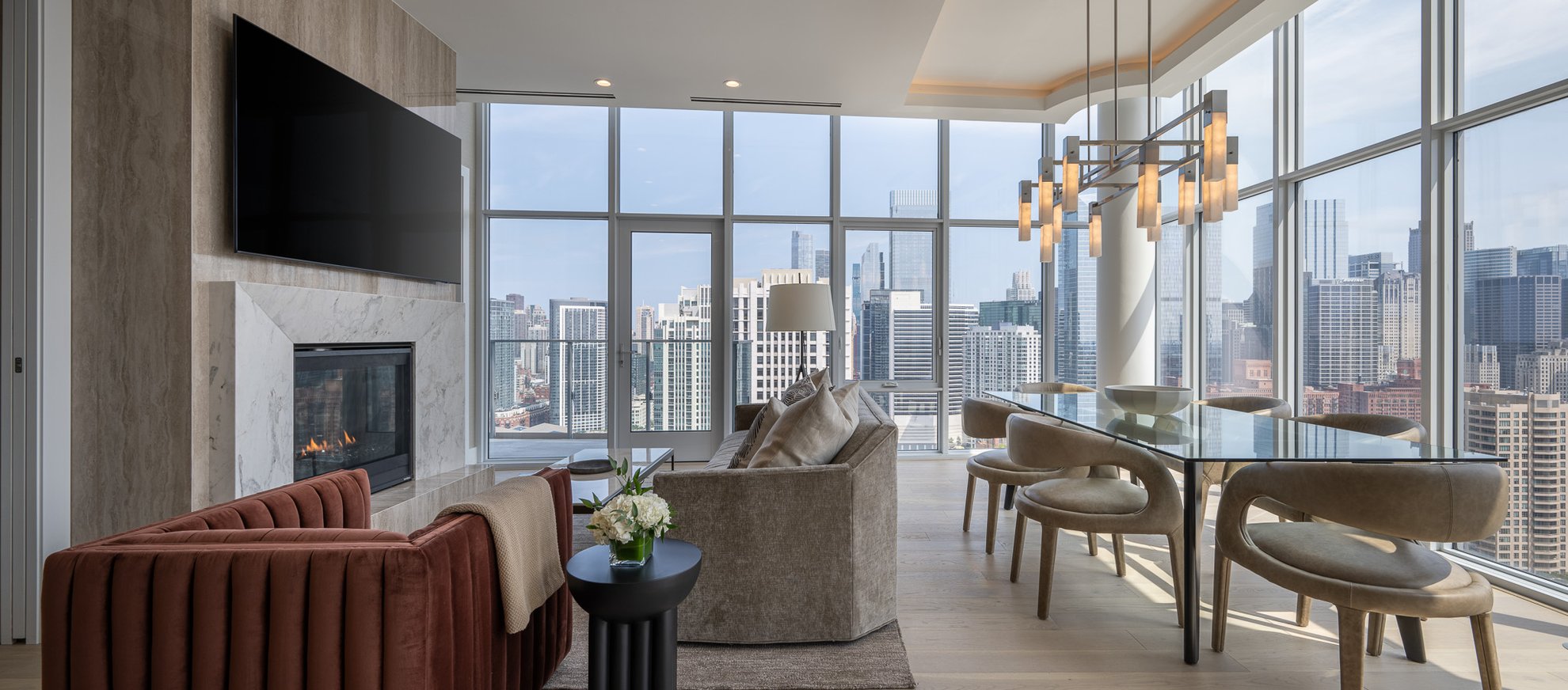 Modern living and dining area with floor-to-ceiling windows offering a city skyline view, featuring a fireplace, wall-mounted TV, velvet seating, glass dining table, and contemporary chandelier.