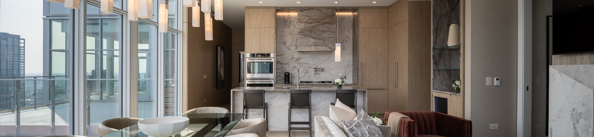 Modern open-concept kitchen and living area with marble accents, wooden cabinets, a dining table, barstools at the island, large windows, and a view of the city skyline. Contemporary pendant lights hang from the ceiling.