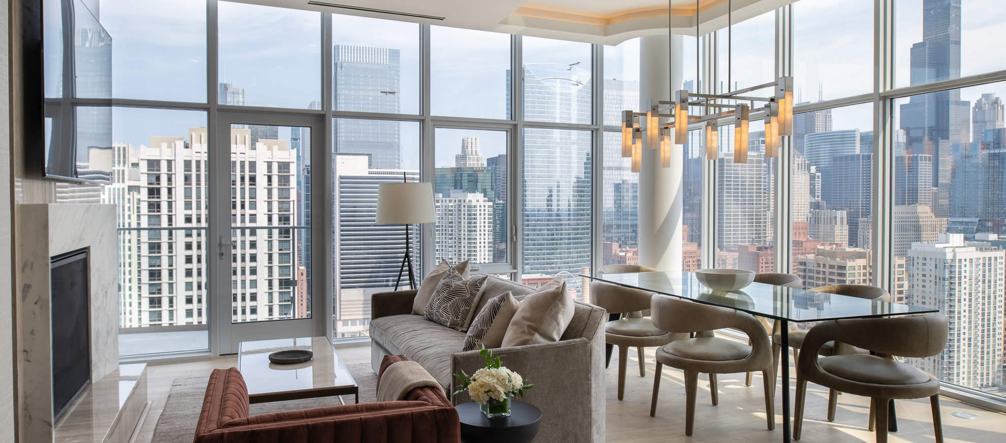 Modern high-rise apartment with floor-to-ceiling windows, a sofa, armchair, dining table, and city skyline views. Contemporary lighting fixture hangs above the table. Bright, airy, and elegantly furnished space.
