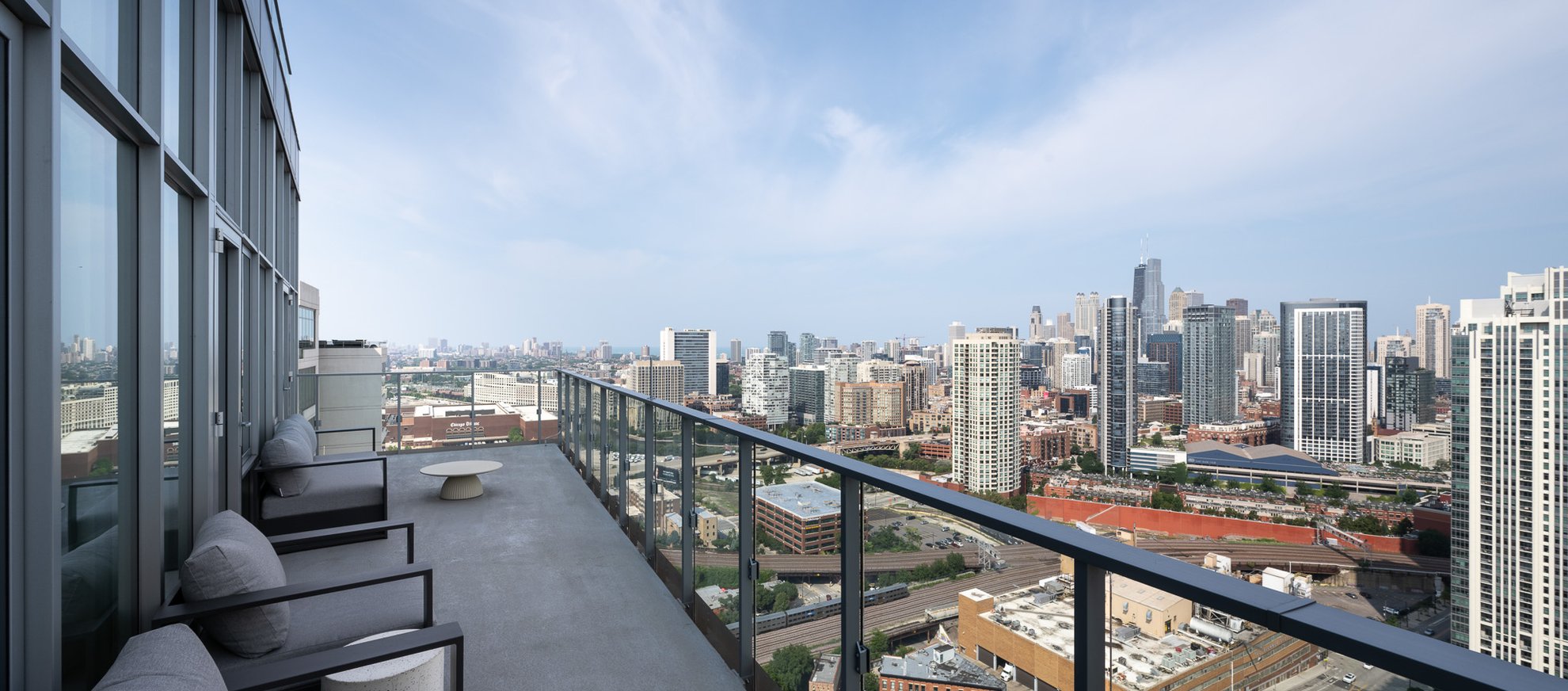 A modern high-rise balcony with glass railing, outdoor seating, and a table overlooks a city skyline filled with tall buildings under a clear blue sky.