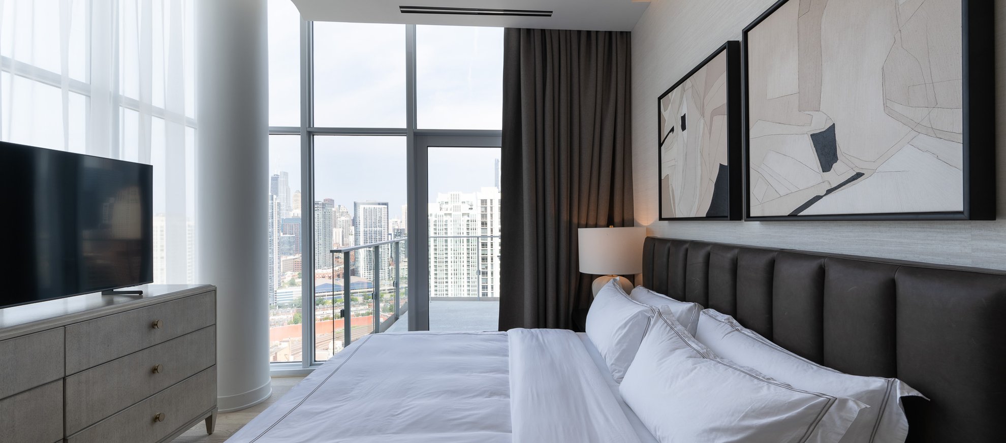 A modern bedroom with a large bed, white bedding, abstract art on the wall, a flat-screen TV on a dresser, and floor-to-ceiling windows showing a city skyline view.