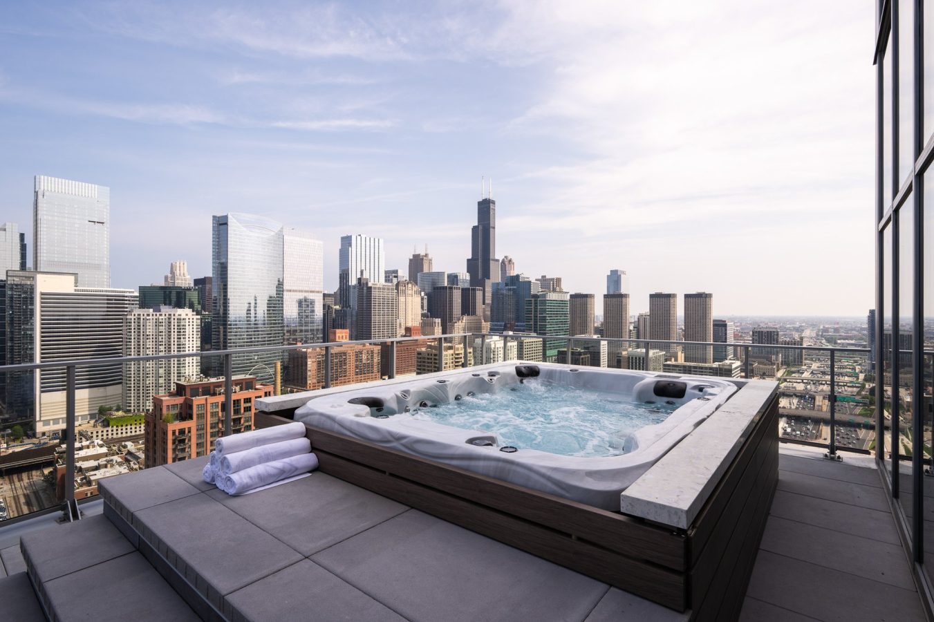 A rooftop hot tub with bubbling water and folded towels overlooks a city skyline with modern skyscrapers under a partly cloudy sky.