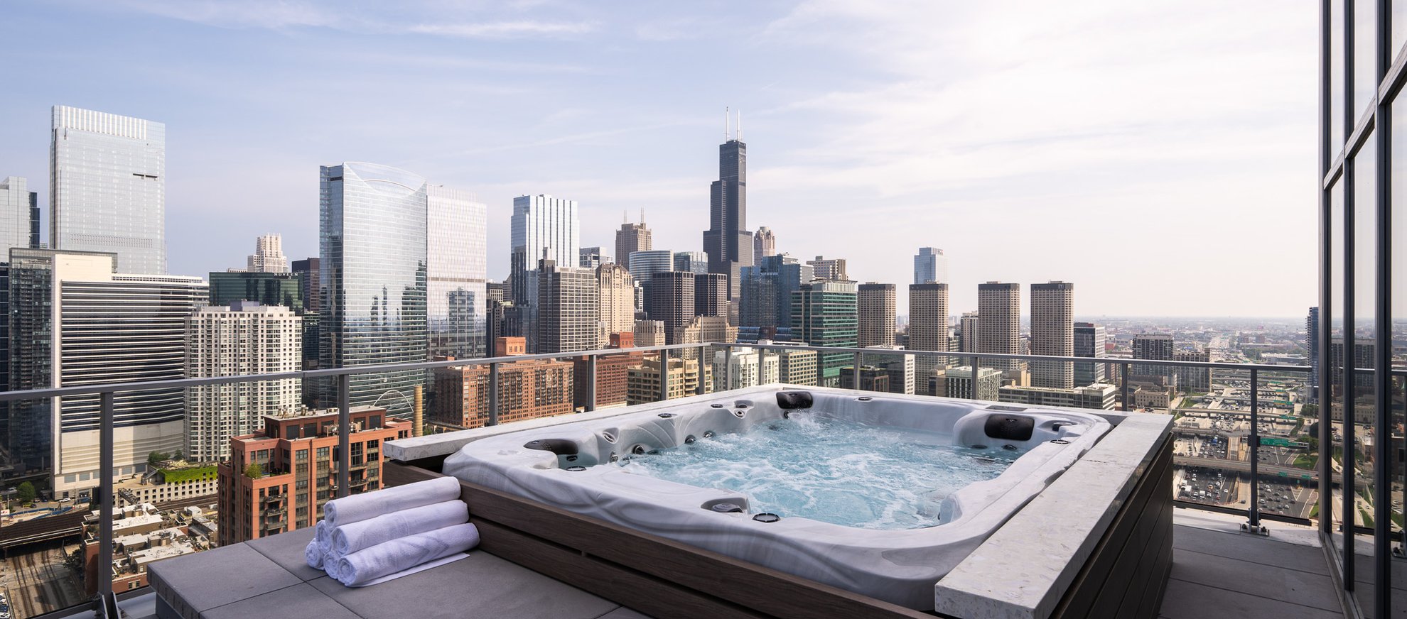 A rooftop hot tub with bubbling water and two rolled white towels overlooks a city skyline filled with modern skyscrapers under a partly cloudy sky.
