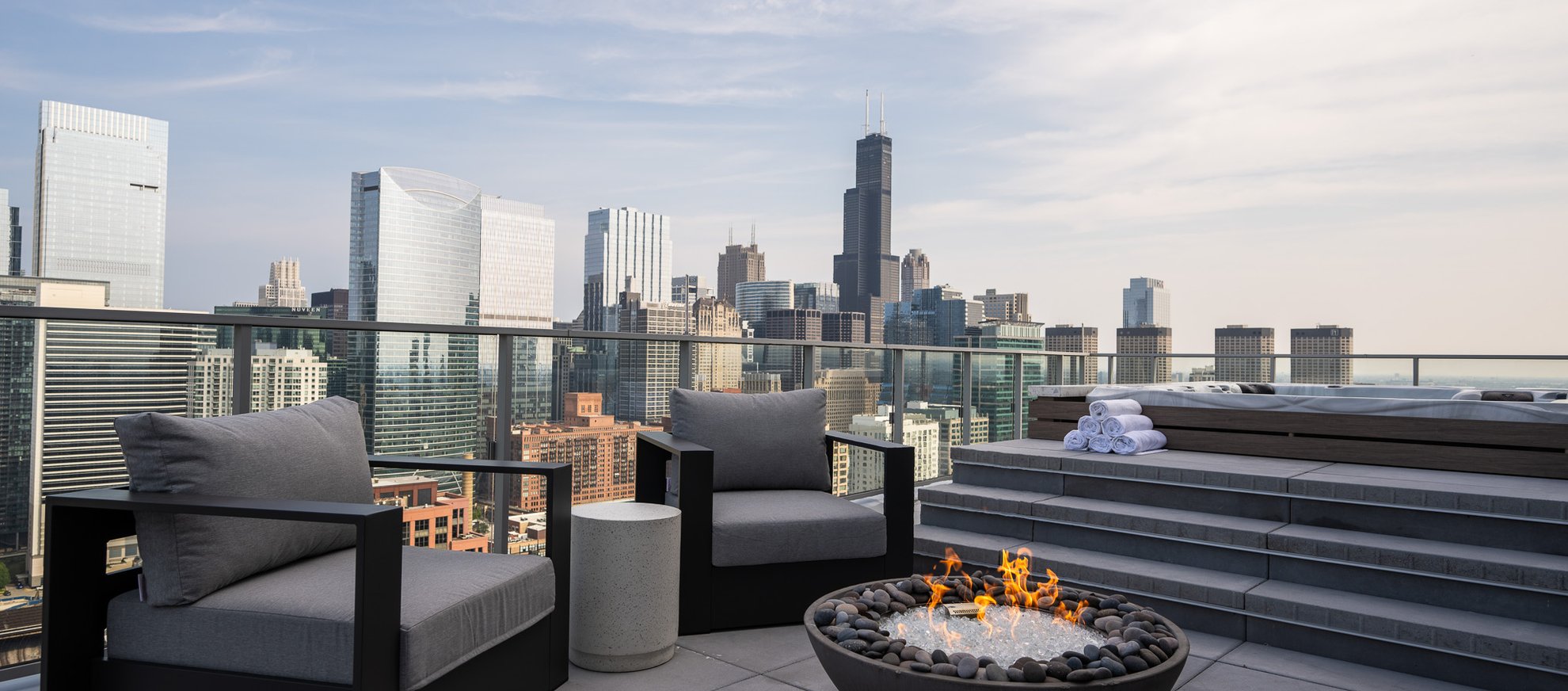 A modern rooftop patio with cushioned chairs, a round fire pit, a stack of towels, and a hot tub, overlooking a city skyline with high-rise buildings under a partly cloudy sky.