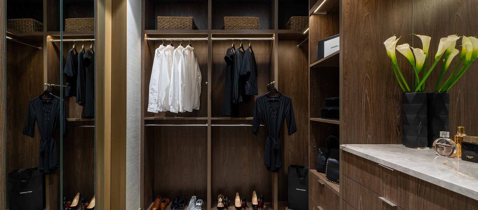 A modern walk-in closet with wooden shelves and cabinets, neatly organized with hanging clothes, shoes, baskets, handbags, and a countertop holding a vase of calla lilies and perfume bottles.
