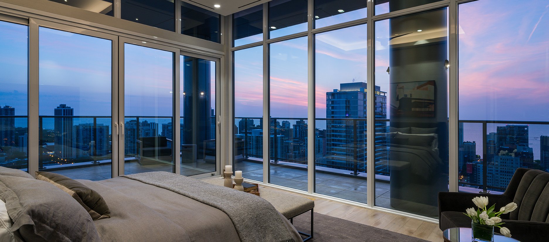 Modern bedroom with floor-to-ceiling windows and balcony overlooking a city skyline at sunset, featuring a large bed, soft lighting, and contemporary decor.