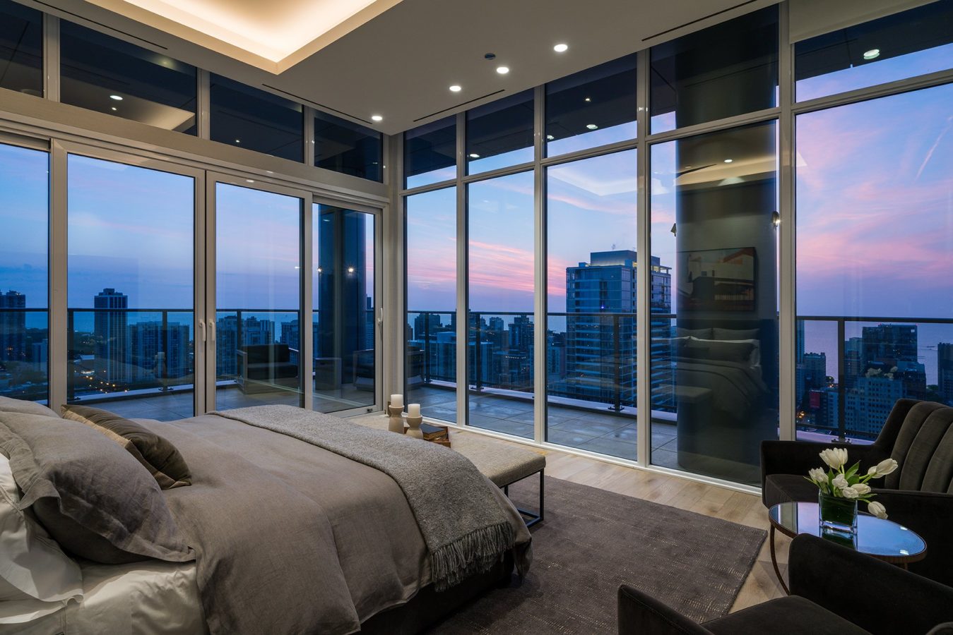 Modern bedroom with floor-to-ceiling windows showing a cityscape at sunset, a neatly made bed with gray bedding, a seating area, and access to a balcony with glass railing.