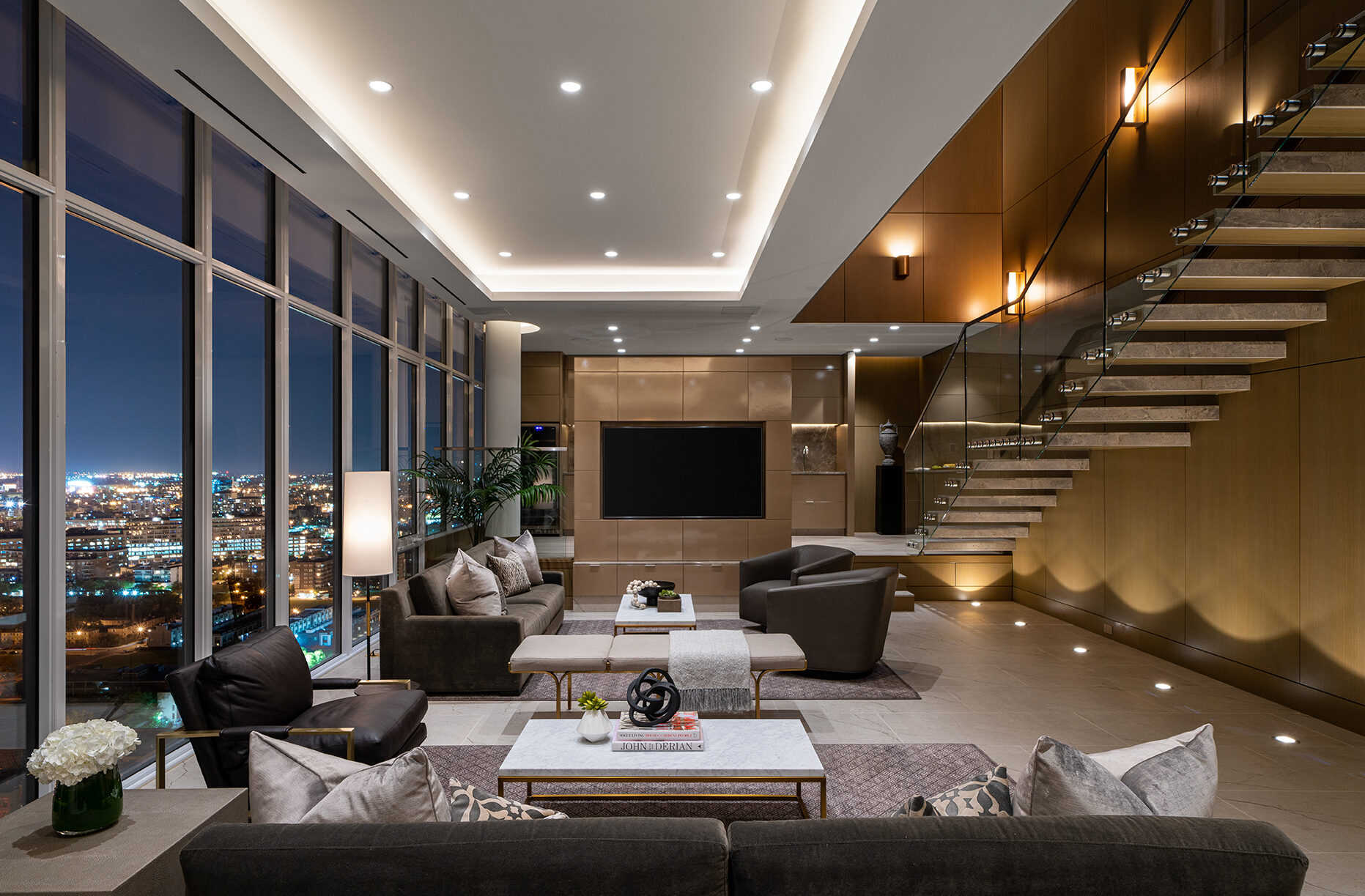 Modern, luxurious living room at night with large floor-to-ceiling windows showcasing a city view, stylish furniture, neutral colors, recessed lighting, and a floating staircase with glass railing along a wood-paneled wall.