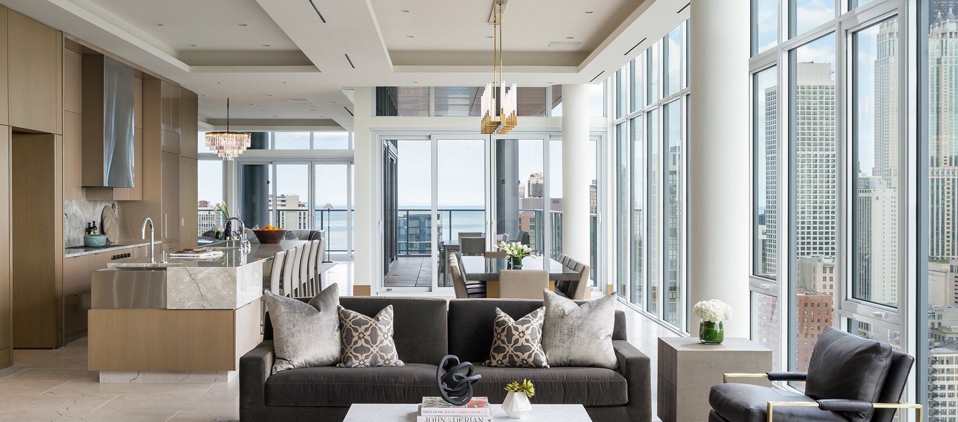 A modern, luxurious open-plan living space with floor-to-ceiling windows, gray sofas, a marble kitchen island, dining area, and city and lake views visible outside. Light colors and contemporary decor throughout.
