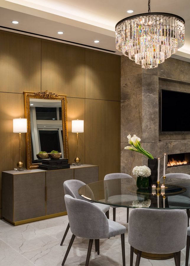 Modern dining room with a glass table, gray velvet chairs, a marble fireplace, elegant chandelier, and a sideboard with a gold-framed mirror, two lamps, and floral decor. Warm lighting creates a cozy atmosphere.