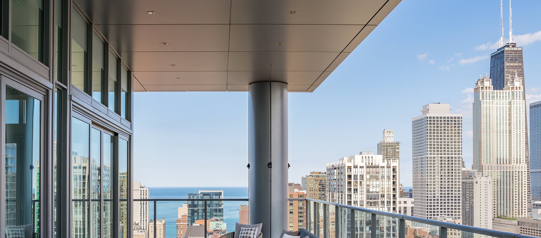 A modern high-rise balcony with glass railings and a view of city skyscrapers and Lake Michigan under a clear blue sky.