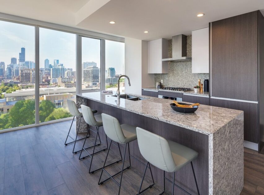 Modern kitchen with a large granite island, four light gray bar stools, built-in appliances, and floor-to-ceiling windows offering a city skyline view in the background.