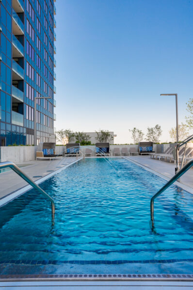 A modern outdoor swimming pool with clear blue water, surrounded by poolside seating and railings, located next to a tall glass building under a clear sky.